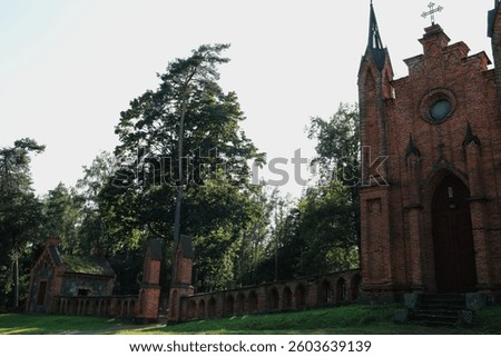 Historic brick chapel with arched windows and tall spires stands beside a cemetery wall and gate, surrounded by dense green forest. The architecture features gothic elements and weathered textures