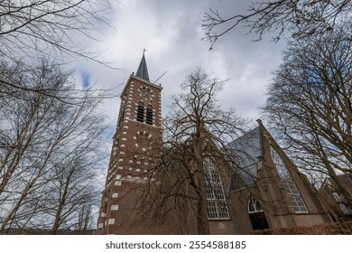 A historic brick church surrounded by tall, bare trees with visible roots. The gothic architecture is highlighted against an overcast sky, creating a serene atmosphere. Church near Amsterdam. - Powered by Shutterstock