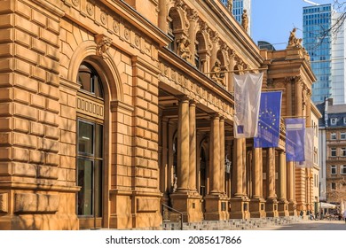 historic building of the stock exchange in Frankfurt with flags above the entrance. brown colored commercial building in baroque style. Skyscrapers in the background. Trees in the foreground in spring