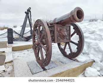 A historic cannon stands prominently next to an anchor, surrounded by snow and showcasing the winter landscape of a coastal area. - Powered by Shutterstock