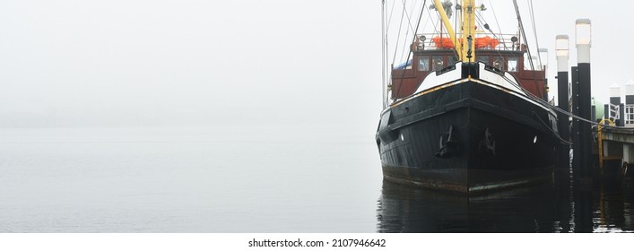 Historic Cargo Boat Moored To A Pier, In A Thick White Morning Fog, Close-up. Kiel, Germany. Transportation, Fishing, Sailing, Logistics, Industry, Traditional Craft, Past, History. Panoramic Image