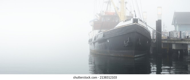 Historic Cargo Boat Moored To A Pier, In A Thick White Morning Fog, Close-up. Kiel, Germany. Transportation, Fishing, Sailing, Logistics, Industry, Traditional Craft, Past, History. Panoramic Image