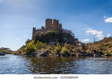 A historic castle stands majestically atop a rocky island surrounded by calm river waters, with a backdrop of lush hills under a clear, bright sky - Powered by Shutterstock