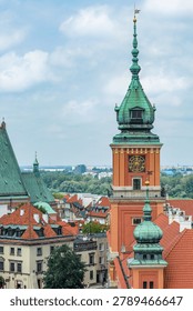 Historic clock tower and red rooftops in Warsaw Old Town, Poland, with copper spires under a cloudy sky.