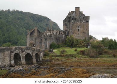 Historic Eilean Donan Castle in the Scottish Highlands, with its stone bridge crossing over a rocky shoreline, surrounded by lush greenery and misty hills in the background. - Powered by Shutterstock