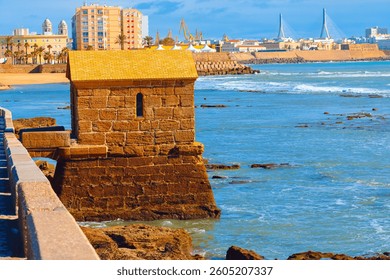 Historic fort stands at the edge of ocean in Cadiz, Spain, blending with rugged coastline. In background, modern buildings, sandy beach and striking cable-stayed bridge contrast with city heritage