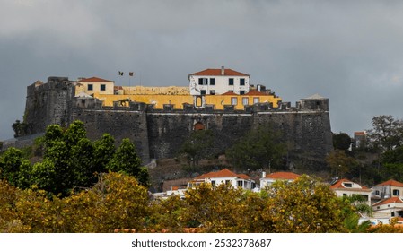 A historic fortress perched on a hilltop, surrounded by trees and houses, with flags flying atop, under a cloudy sky, showcasing its commanding presence - Powered by Shutterstock