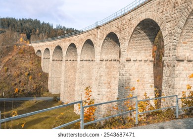 The historic railway viaduct in Smrzovka stands majestically under the clear sky, framed by autumn foliage in the Jizera Mountains, showcasing vibrant colors along the tranquil landscape. - Powered by Shutterstock