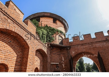 A historic red brick castle structure features a round tower partially covered in green ivy, with arched gateways and crenellated battlements against a bright sky