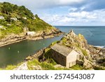 A historic stone building sits atop Polperro’s rugged cliffs, overlooking the calm Cornish sea under a bright, clouded sky.