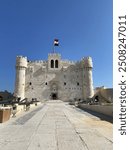 Historic stone fortress with crenellated towers, arched entrance, and Egypt flag, viewed through an archway against a clear blue sky. Located at the Citadel of Qaitbay, Alexandria, Egypt