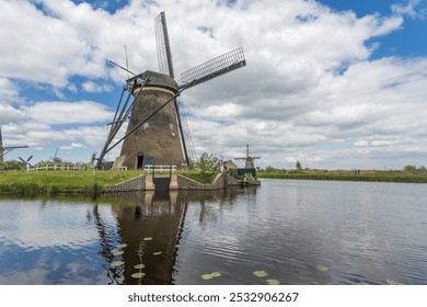 Historic windmill stands gracefully by a calm waterway, its reflection mirrored. Lush grass and a partly cloudy sky enhance the idyllic scene. - Powered by Shutterstock