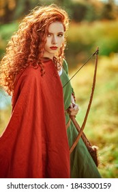 Historical reconstruction. Beautiful young woman archer with magnificent long red hair in a historical celtic dress stands on the edge of the forest.