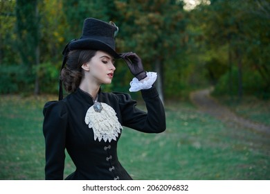 Historical reconstruction of the late 19th and early 20th century. An elegant dark-haired lady in a strict black dress and hat walks through the park. Historical makeup and hairstyle.