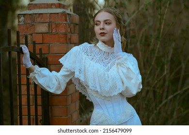 Historical Reconstruction Of The Victorian Era. Portrait Of A Beautiful Noble Lady In A White Dress Standing By The Gate Of A House. Hair And Makeup, Late 19th Century.