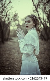 Historical Reconstruction Of The Victorian Era. Portrait Of A Beautiful Noble Lady In A White Dress Walking In An Old Park. Hairstyle And Makeup, Late 19th Century. Fashion.