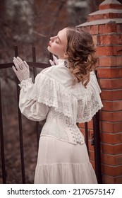 Historical Reconstruction Of The Victorian Era. Portrait Of A Beautiful Noble Lady In A White Dress Standing At The Palace Fence. Hairstyle And Makeup, Late 19th Century.