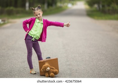 Hitch-hiking little funny girl on the road with a suitcase and a Teddy bear.
