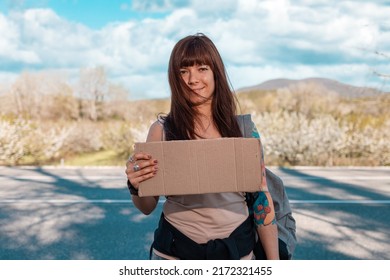 Hitchhiking. Portrait of smiling caucasian young woman with tattoed hand holding a cardboard sign with mock up. Copy space. The concept of local traveling.