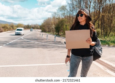 Hitchhiking. Pretty young woman in sunglasses and cap holding a cardboard sign. Mck up. Empty road on the background. The concept of local traveling.