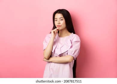 Hmm, Interesting. Pensive And Cunning Asian Girl Having Idea, Plan To Do Something, Looking At Camera And Touching Chin With Thoughtful Face, Standing On Pink Background