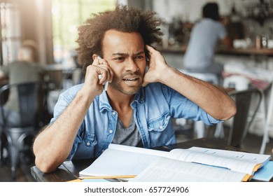 Hnadsome Dark-skinned College Student Dressed Casually Looking Frustrated, Plugging One Ear While Trying Hard To Hear What They Are Saying On The Other End Of Line, Asking What? Come Again Please?