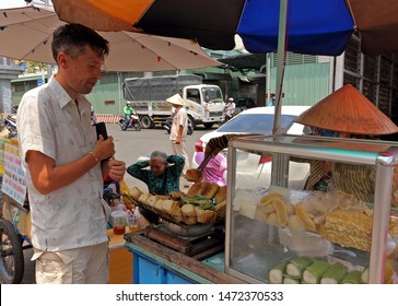 Ho Chi Minh City, Vietnam - March 28, 2019: European foreigner buys fried sticky rice cake with banana in street stall on market. Tourist gets acquainted with Vietnamese local cuisine