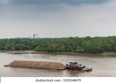 Ho Chi Minh City, Vietnam - March 12, 2019: Song Sai Gon River. Short Tugboat Pushes Long Barge Filled With A Long Mount Of Sand. Brown Water, Green Belt Of Jungle, And Light Blue Sky.