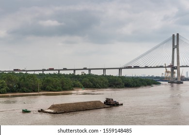 Ho Chi Minh City, Vietnam - March 12, 2019: Song Sai Gon River. Short Tugboat Pushes Long Barge With Sand. Phu My Bridge In Back. Brown Water, Green Belt Of Jungle, And Light Blue Sky.