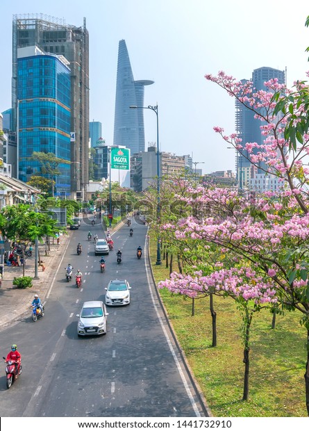 Ho Chi Minh city, Vietnam, March 16th, 2019:\
Traffic in Saigon from high view, street with motorbikes, car move\
under pink tabebuia rosea flower tree of developed city in Ho Chi\
Minh City, Vietnam