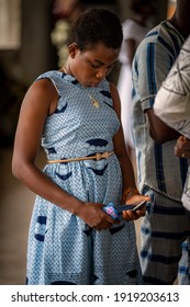 Ho, Ghana - September 16, 2018: An African Teen Girl In A Blue Dress Stands In Church Holding A Cell Phone And Sneaks A Look At The Screen.