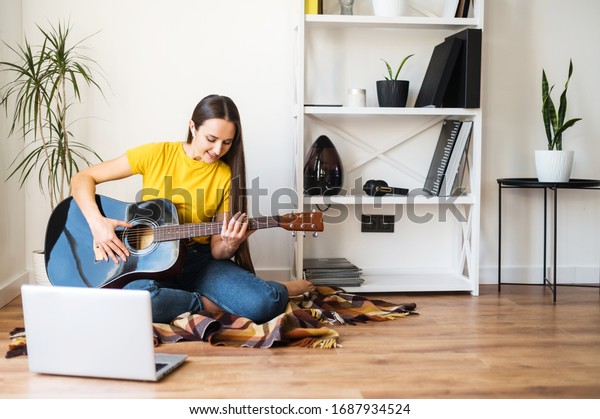 Hobbies\
and leisure activities during quarantine. Online training, online\
classes. A young woman watches a video lesson on playing the\
guitar, she sits on a cozy plaid with a\
guitar