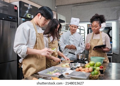 Hobby cuisine course, senior male chef in cook uniform teaches young cooking class students to peel and chop apples, ingredients for pastry foods, fruit pies in restaurant stainless steel kitchen.