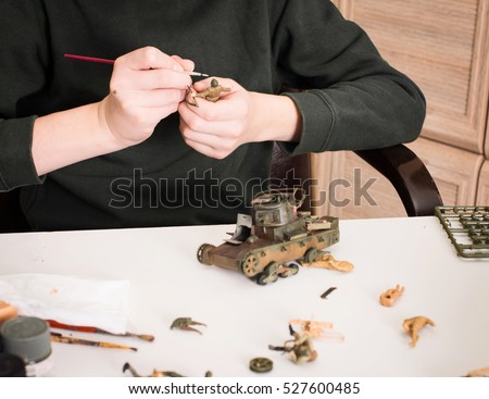 Hobby and leisure concept. Teen boy assembling and painting plastic models of soldiers and tank at workplace in his room. 