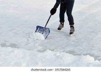 Hockey Player On Ice-skating Removing Snow With A Blue Shovel In The Winter In A Park. Wearing Dark Clothes. Moving Towards Camera. Ice Rink Cleaning At Corner. Active Sport Outdoor