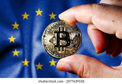 Hold The Physical Version Of Bitcoin (the New Virtual Currency) And The European Union Flag. EU Cryptocurrency And Blockchain Technology Investor Concept Map (the Background Is Deliberately Blurred)