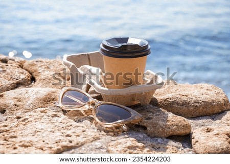 Holder with sunglasses and takeaway cup of hot coffee on beach