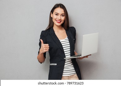 Holding asian business woman holding laptop computer and showing thumb up while looking at the camera over gray background