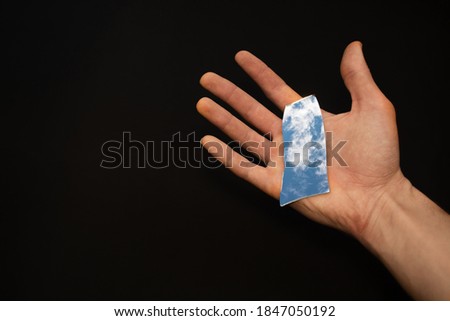 Holding blank broken mirror splinter in palm of hand against black background which reflects blue sky with white clouds. Copy space. Top view 