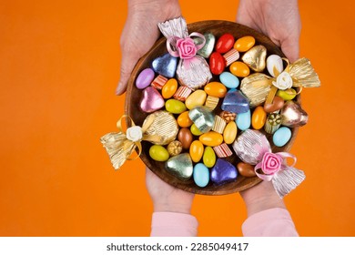 Holding bowl of sweet, woman and child hands holding bowl of sweet. Top view image of wrapped luxury chocolate and almond dragee in wooden tray. Isolated orange studio background with copy space.