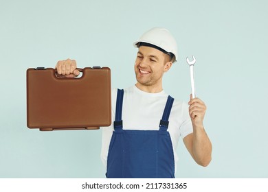 Holding Brown Case. Worker In Uniform And Hard Hat Standing In The Studio With Equipment.