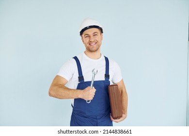 Holding Brown Case. Worker In Uniform And Hard Hat Standing In The Studio With Equipment.