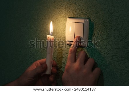 Holding burning candle in a dark room near a light switch, blackout. 
Power outage, energy crisis. Problems with power supply. Male finger turning on and off the light switch at night
