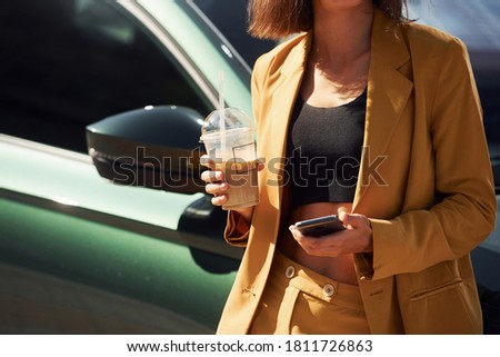 Holding drink and smartphone. Young fashionable woman in burgundy colored coat at daytime with her car.