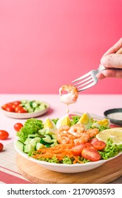 Holding Fork With Prawn. Healthy Salad On Wood Cutting Broad Ingredients: Fresh Green Coral Lettuce, Prawn, Cherry Tomato, Cucumber, Carrot, Lemon And Egg In A White Plate On Pink Background