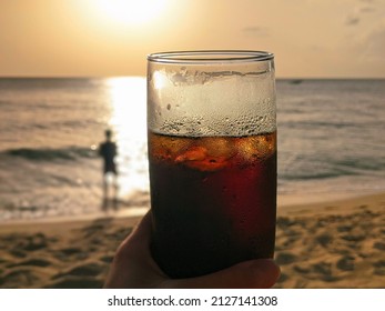 Holding A Glass Of Rum And Coke With Ice With The Beach At A Golden Sunset In The Background