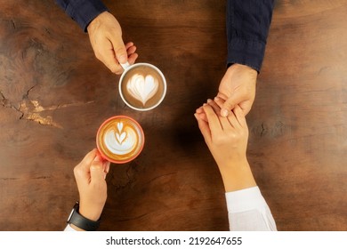 Holding hands, top view photo of young lovers holding hands. Drinking coffee with hearth shape latte art. Sitting on wooden table. Close up man and woman hands, love story concept idea background. - Powered by Shutterstock