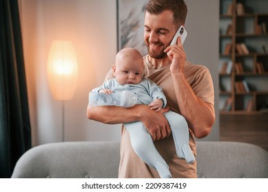 Holding Kid And Talking By Phone. Father With His Newborn Baby Is Indoors. Conception Of Single Dad.