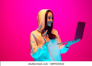 Holding laptop, videocall. Caucasian woman's portrait on pink studio background in mixed neon light. Student, worker. Concept of human emotions, facial expression, sales, ad, fashion. Copyspace.