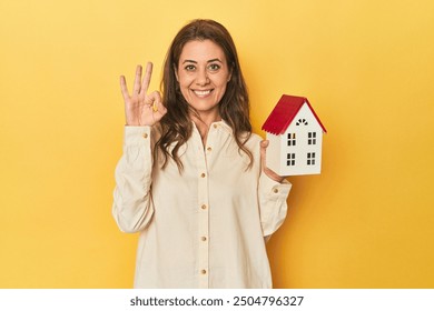 Holding miniature house, realty concept cheerful and confident showing ok gesture. - Powered by Shutterstock